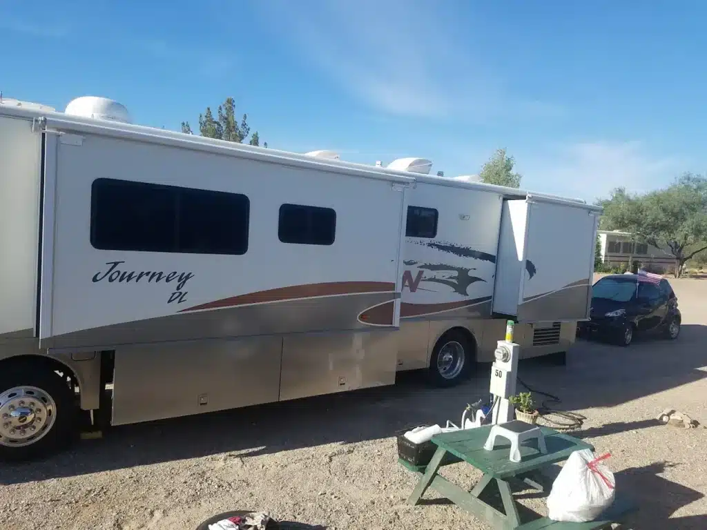 A white Winnebago Journey DL motorhome with slide-outs parked at a gravel RV site with an electric hookup pedestal