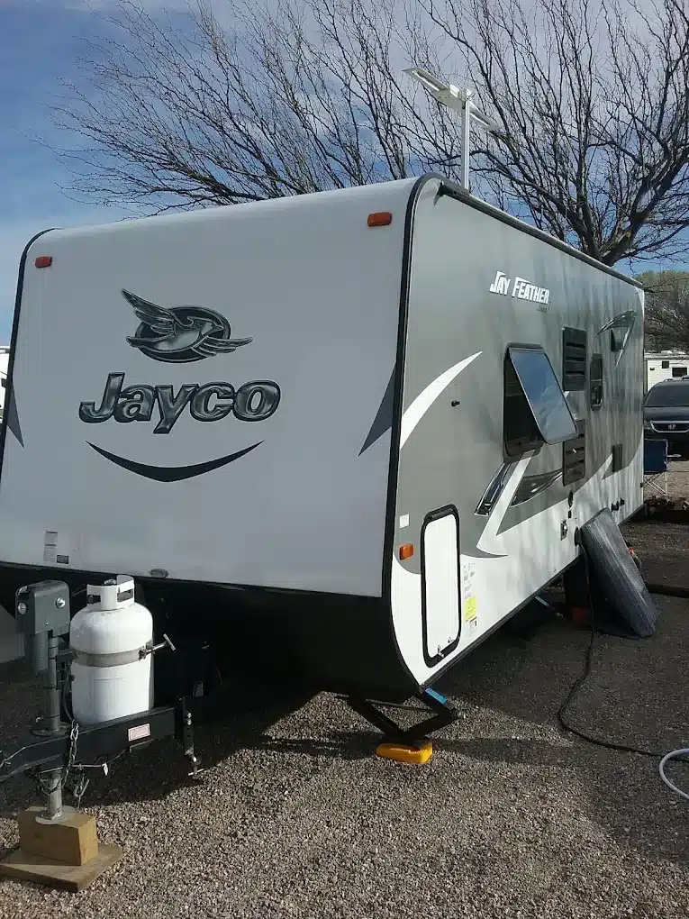A white Jayco Jay Feather travel trailer parked on gravel with a propane tank, stabilizer jacks, and bare trees behind.